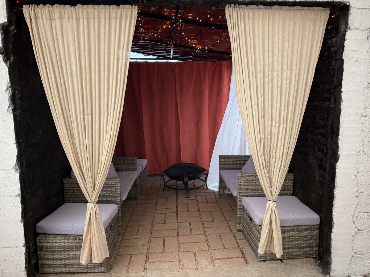 An outdoor sitting area is enclosed by soft curtains, creating a cozy retreat. Two wicker seating arrangements with cushions are positioned around a central fire pit. String lights hang above, adding a subtle glow to the space, while the backdrop features decorative fabric in warm tones.