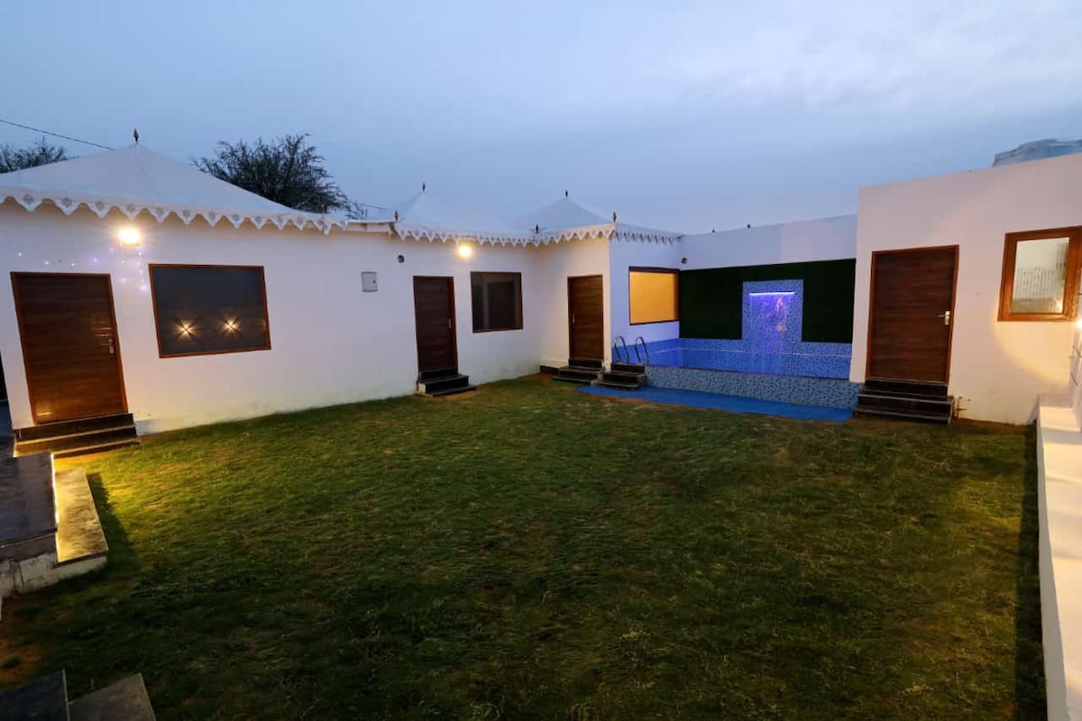 A tranquil outdoor area is framed by several private entrances to tent domes. Soft ambient lighting enhances the spacious lawn, alongside a blue-tiled feature with water flowing gently. The space invites relaxation amidst a peaceful natural setting.
