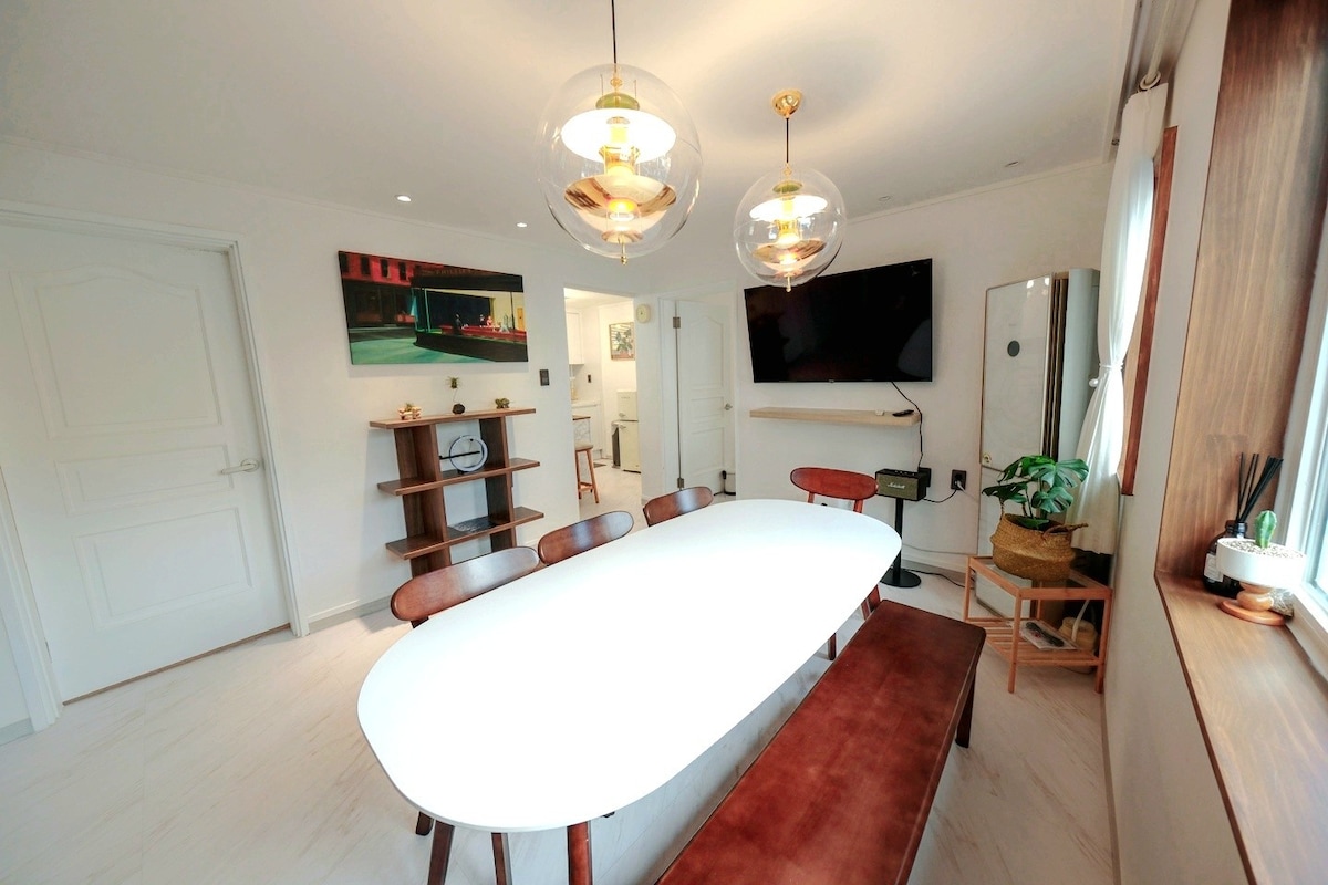 A modern dining area features a spacious white table surrounded by wooden chairs and benches. Soft pendant lights illuminate the space, creating a welcoming atmosphere. A large TV is mounted on the wall, and potted greenery adds a touch of nature near the windows.