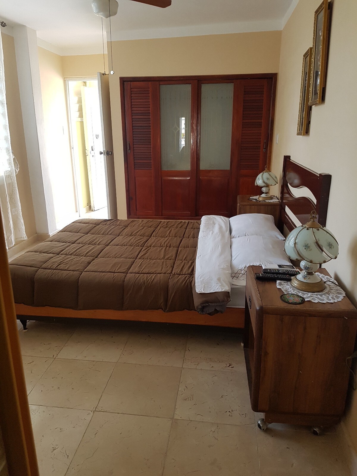 A cozy bedroom features a spacious bed with a brown comforter and white pillows. A wooden nightstand holds a decorative lamp and a remote control. A large wardrobe with sliding doors is visible alongside a partially open door leading to another area.