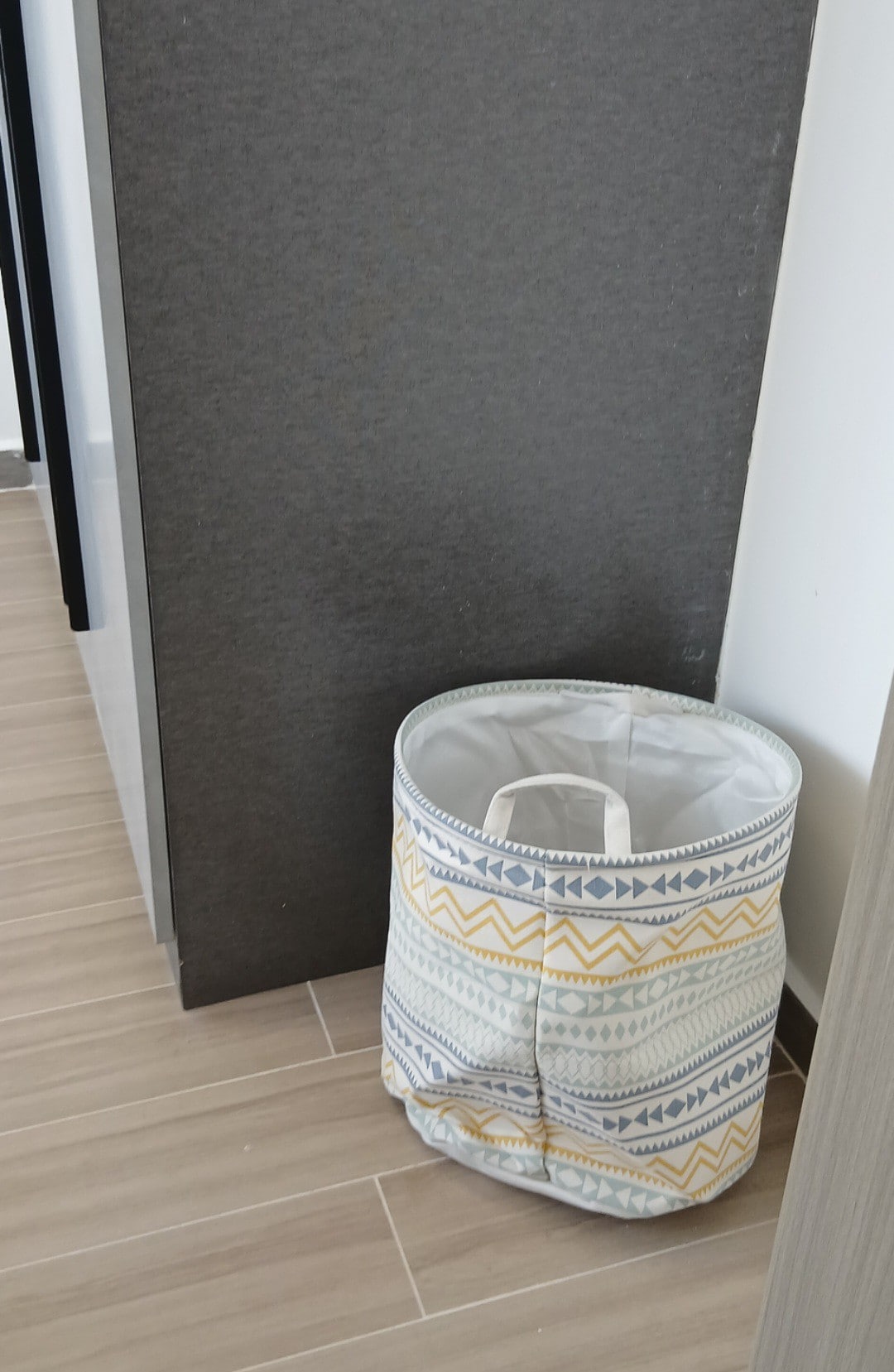 A decorative storage basket with a geometric pattern in muted colors is positioned against a wall. The basket's fabric is complemented by a sturdy white liner and handles, offering practical storage solutions in the space.
