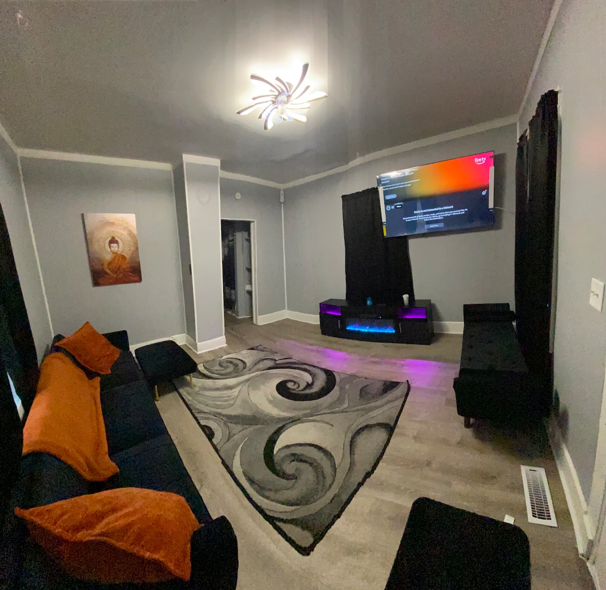 A spacious living room is highlighted by a large dark sofa adorned with orange pillows. A decorative area rug with swirling patterns covers the floor. A sleek entertainment unit with ambient lighting is positioned under a wall-mounted television, while dark curtains flank the large windows.
