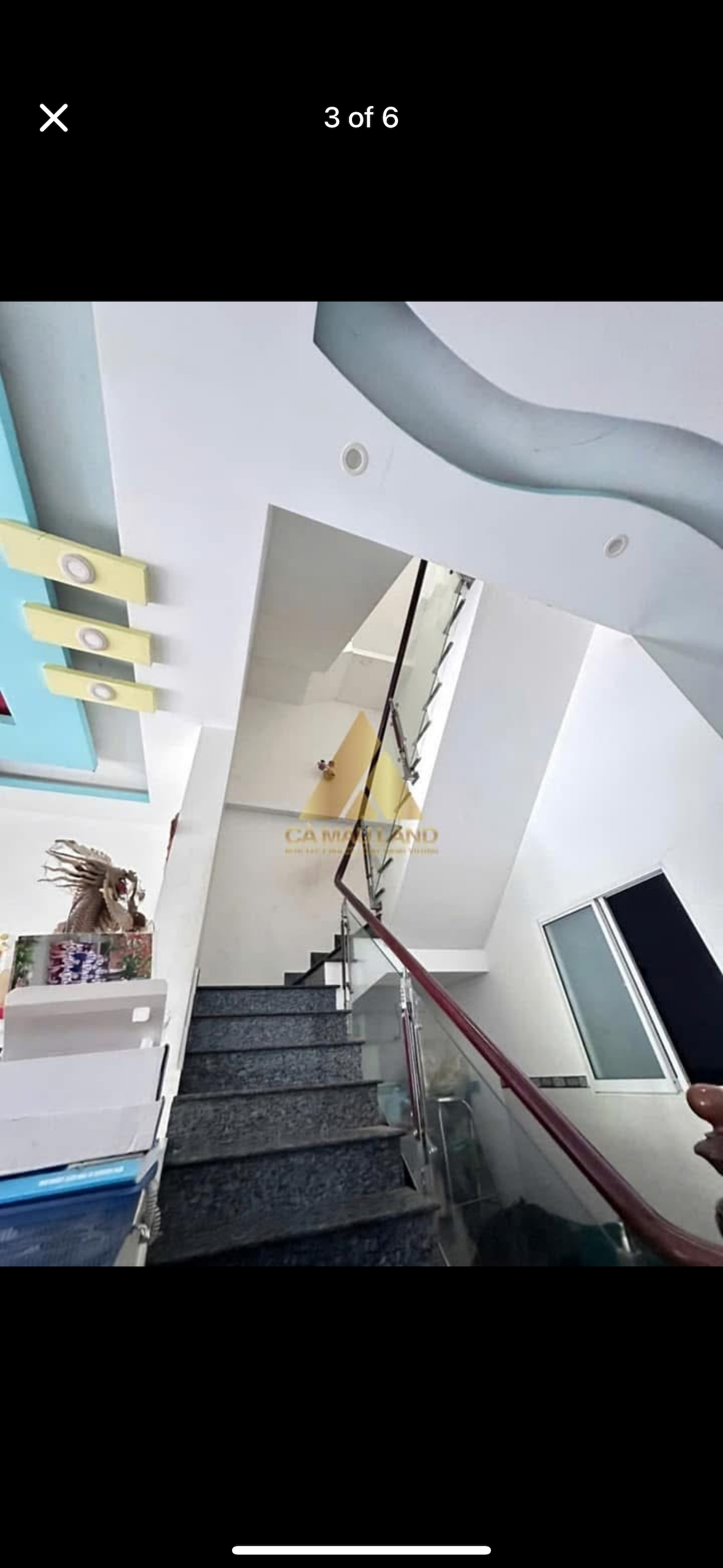 A well-lit staircase is visible, featuring a modern design with glass railings. The walls are adorned with colorful accents, and natural light filters through a nearby window, creating a bright atmosphere. Steps are covered in a dark material, leading upwards.