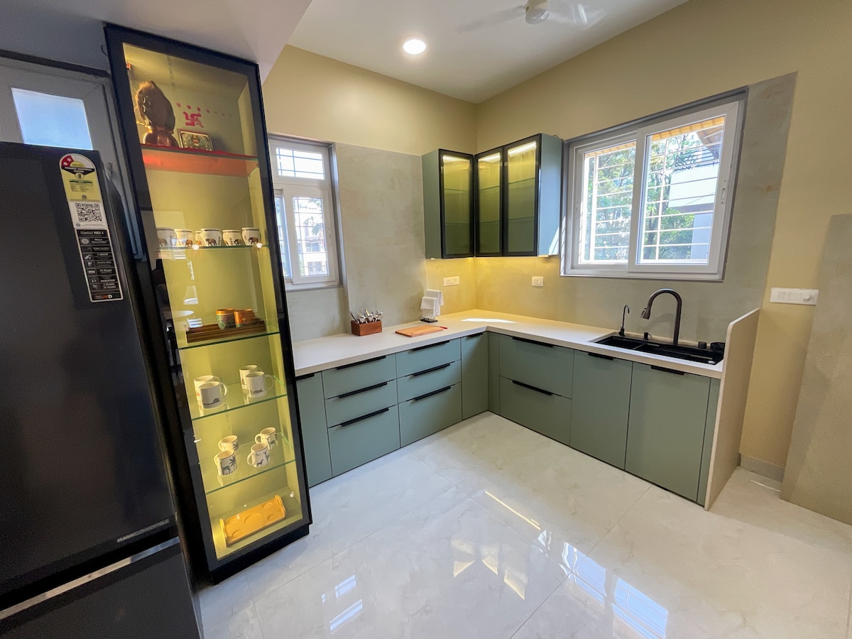 The kitchen features modern cabinetry in subdued tones. Stainless steel appliances are visible alongside a glass-fronted display showcasing dishware. A spacious countertop offers ample preparation space, while a sink is positioned beneath a window that allows natural light to brighten the area.
