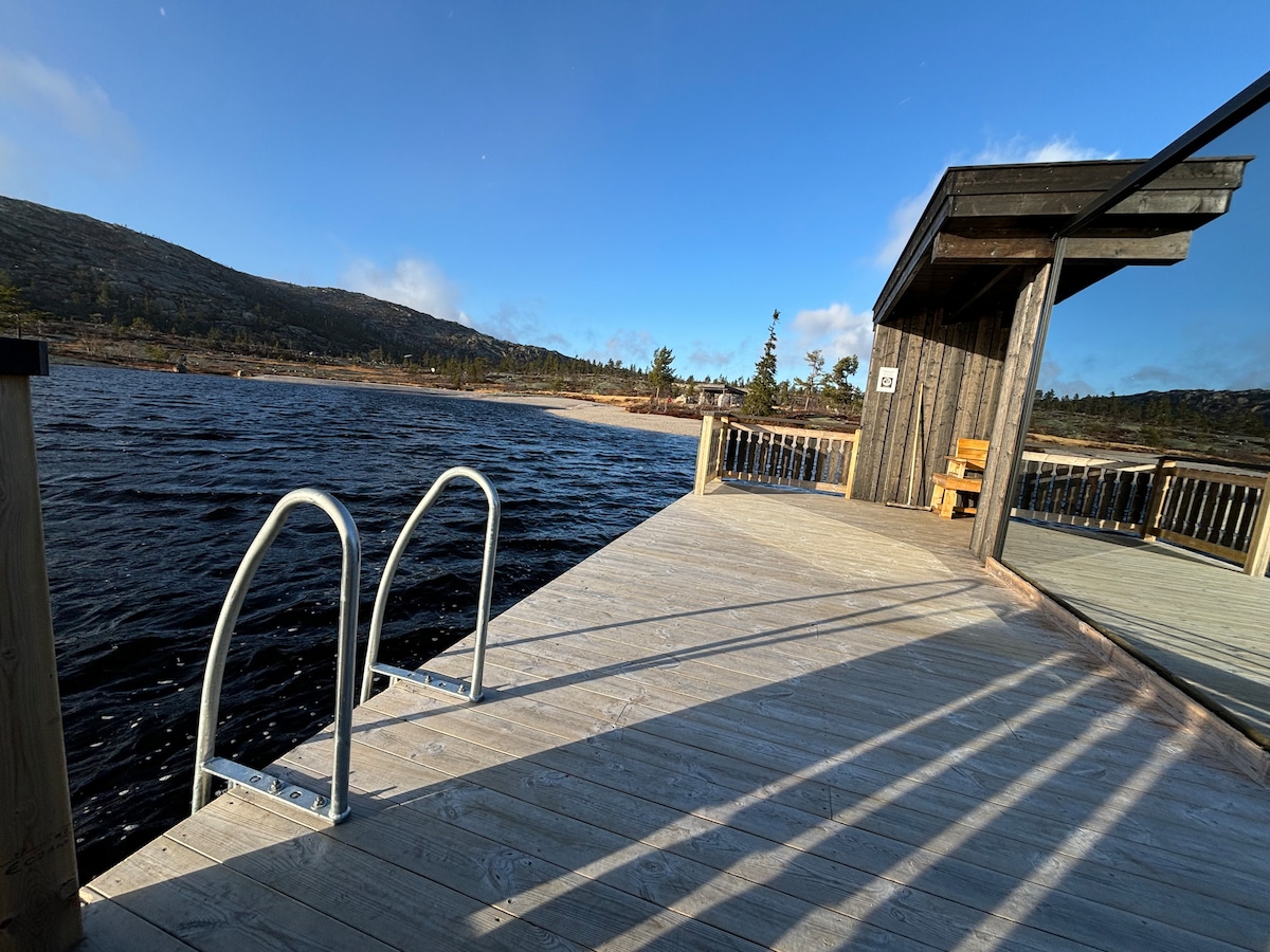 A wooden dock extends over the water, with a set of metal steps leading into it. A small cabin is visible, featuring large windows and a seating area. Surrounding the dock are scenic views of hills and trees under a clear blue sky.