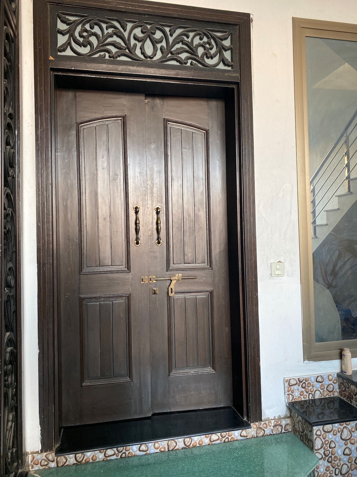 The image displays a pair of elegant wooden doors featuring vertical panels and decorative carvings at the top. A brass handle is positioned on each door, with a sturdy padlock visible below. A tiled entrance area leads up to the doors, enhancing the welcoming entrance.