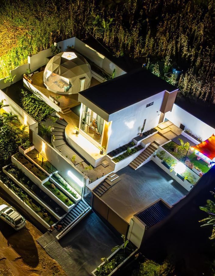 An aerial view captures a modern architectural home featuring a distinct geodesic dome. Surrounding greenery is visible, along with pathways that lead to various outdoor spaces. The night scene highlights the property's illuminated features, creating a serene and unique atmosphere.