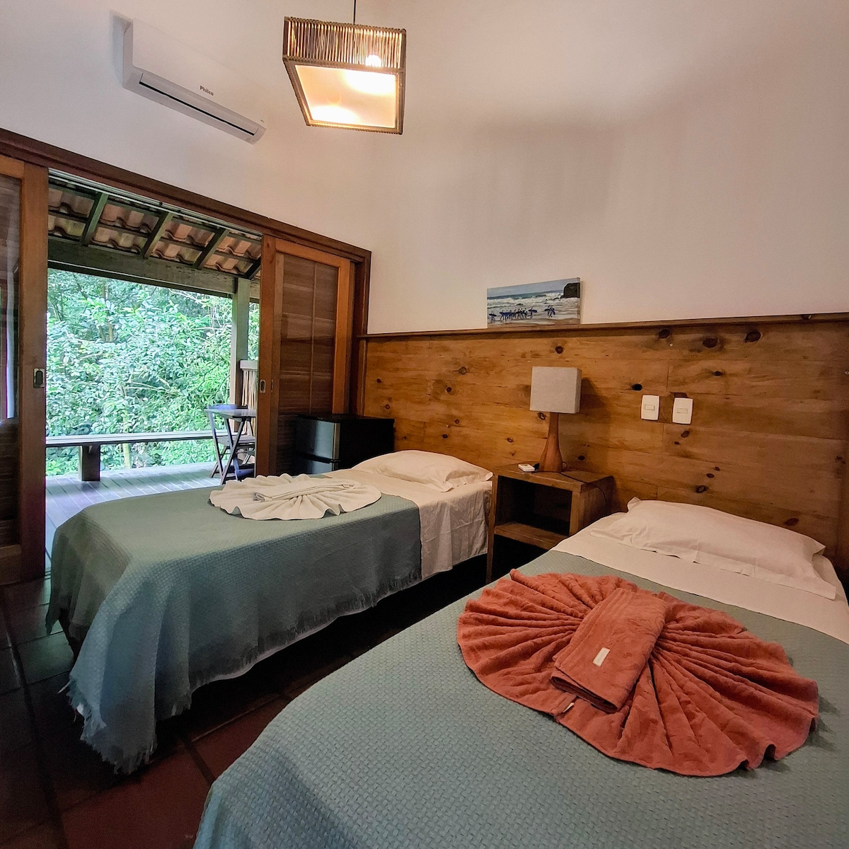 The sleeping area features two comfortable beds covered with soft linens. A natural wood panel accentuates the room, complemented by a bedside lamp. A door leads to a private balcony, where views of the surrounding greenery can be appreciated.