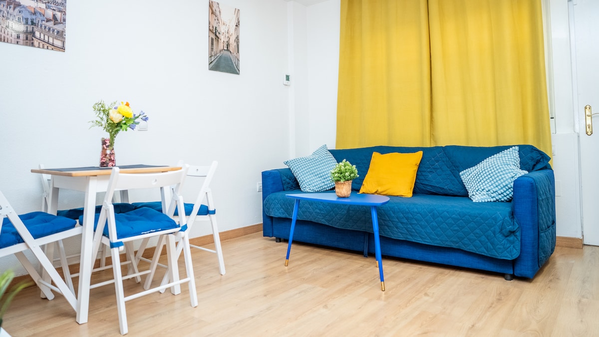 A bright living area features a blue sofa adorned with cushions and a decorative yellow pillow. A small blue table is placed in front of the sofa, accompanied by four white chairs. Light wooden flooring complements the cheerful atmosphere created by the yellow curtains.