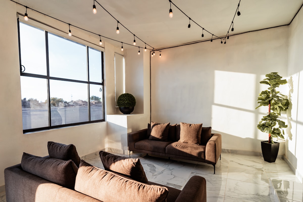 A bright living area features large windows that allow natural light to fill the space. Comfortable brown sofas are arranged thoughtfully on stylish marble flooring, and a potted plant adds a touch of greenery. Soft lighting from hanging bulbs enhances the atmosphere.