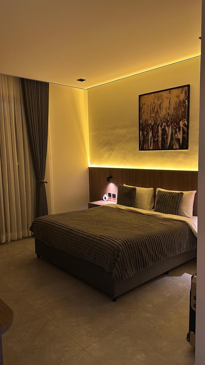 A modern bedroom is illuminated by soft lighting, creating a serene ambiance. A spacious bed with textured bedding is positioned against a softly lit wall. Floor-to-ceiling curtains frame a window, while a framed artwork adds a touch of character to the room.