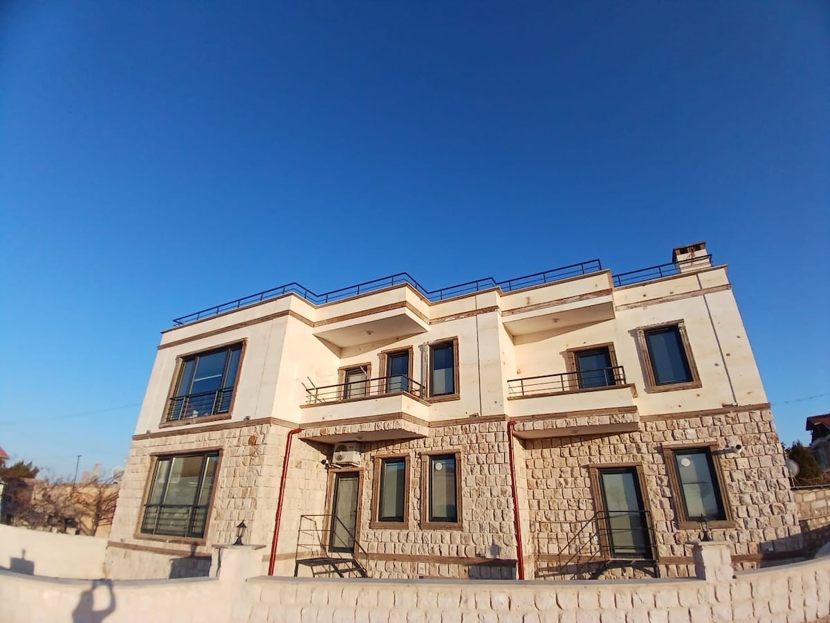 The exterior of a two-story stone building showcases a blend of modern and traditional architectural styles. Large windows are accented with metal railings, and balconies overlook the surrounding area. A clear blue sky provides a bright backdrop to the structure.