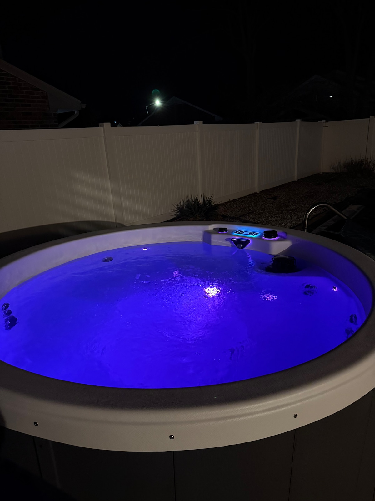 A hot tub is seen illuminated with soft purple lighting, creating a relaxing ambiance. The water appears calm and inviting, surrounded by a white privacy fence and a gravel landscape, providing a secluded atmosphere for enjoyment.