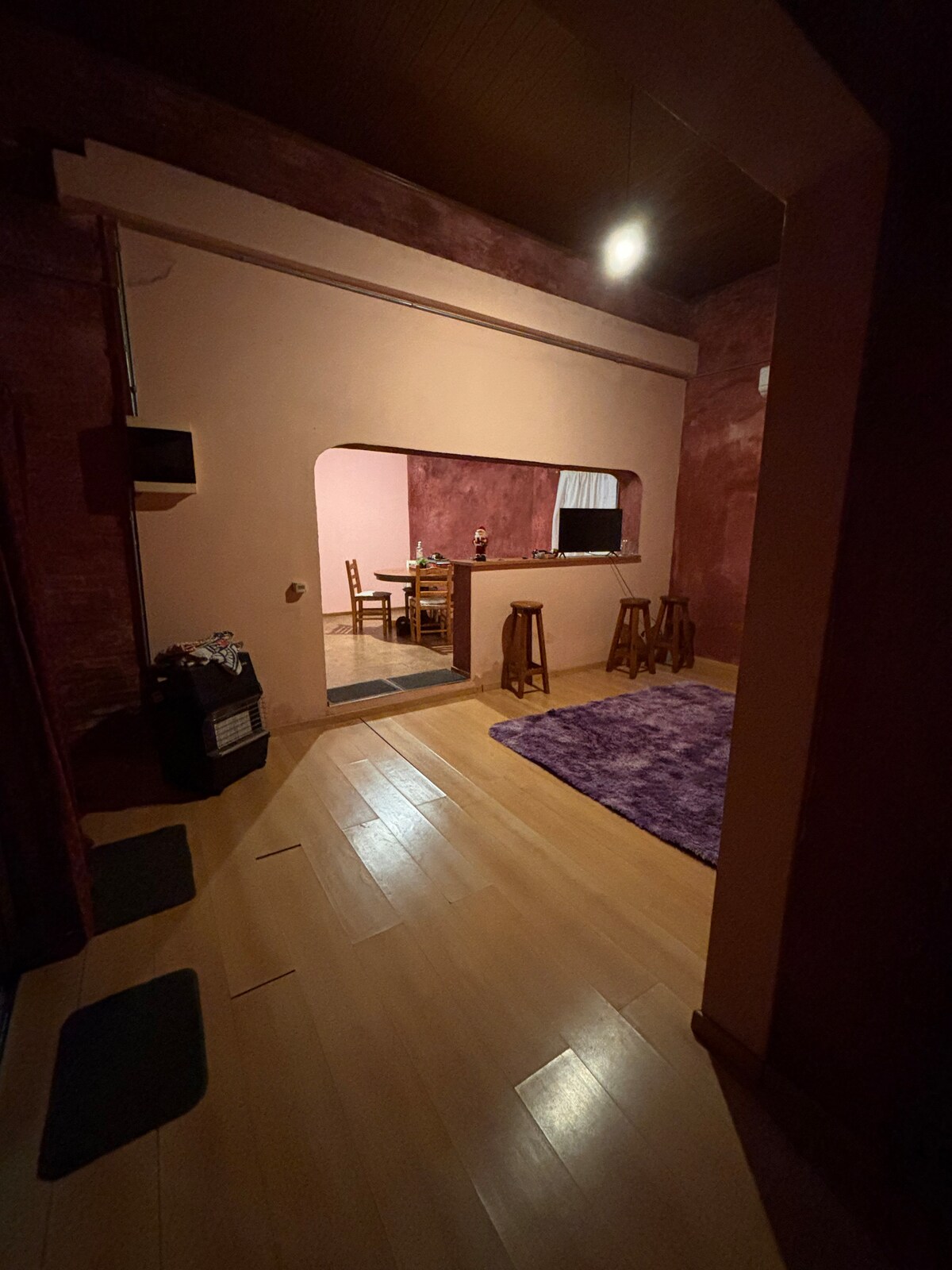 A cozy interior space is featured, with light wooden flooring and a plush purple rug. An arched opening leads to a dining area with a wooden table and chairs. Soft lighting illuminates the room, highlighting textured walls in warm tones.