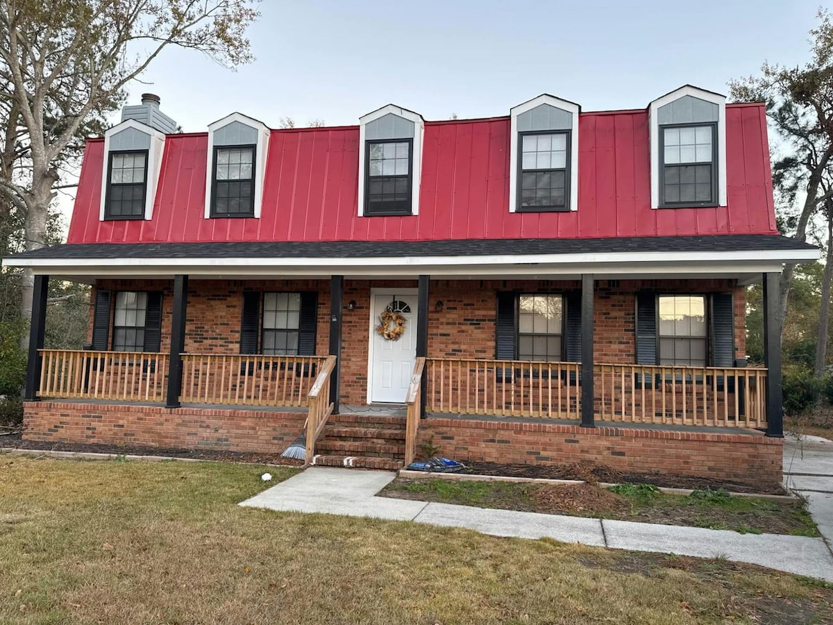 A red-roofed house features a brick façade and a welcoming front porch with wooden railings. Five windows with black shutters illuminate the exterior. Decorative elements, including a wreath, are displayed on the front door, enhancing the home’s inviting appearance.