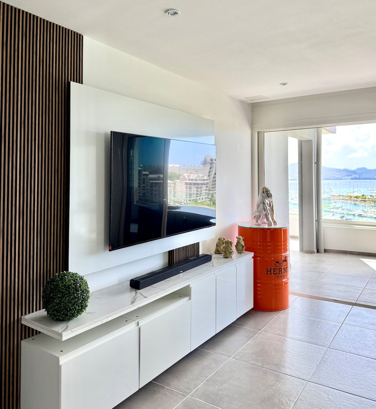A sleek entertainment area features a flat-screen TV mounted on a white wall cabinet, harmonizing with natural wood paneling. A decorative orange cylindrical object adds a pop of color, while large windows reveal views of the marina and allow natural light to fill the space.