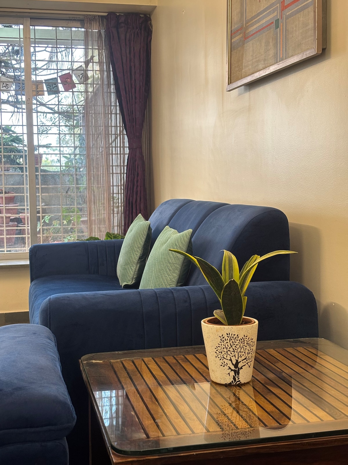 A comfortable blue sofa is paired with two green cushions, positioned next to a tasteful wooden coffee table. A potted plant adds a touch of greenery, while natural light filters in through a window with sheer curtains, revealing a glimpse of the serene backyard.
