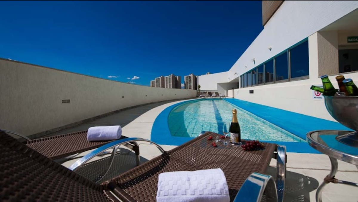 A modern pool area is highlighted by a long, elegantly curved swimming pool surrounded by lounge chairs. A bottle of champagne and a bowl of snacks are placed on a table, with a clear blue sky and distant buildings visible in the background.