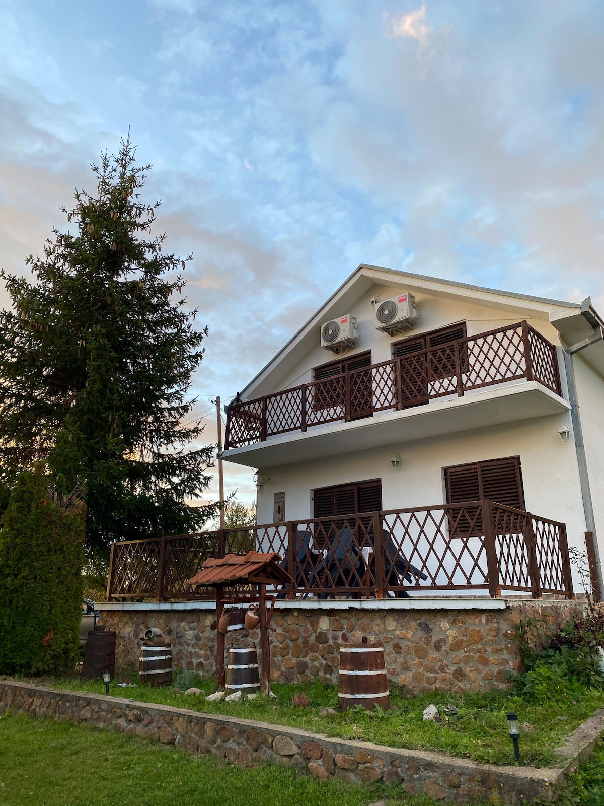 A two-story villa is displayed with a white exterior and brown lattice balconies. Air conditioning units are mounted on the wall. A lush green lawn features a wooden well and decorative barrels, complementing the tranquil outdoor setting under a partly cloudy sky.