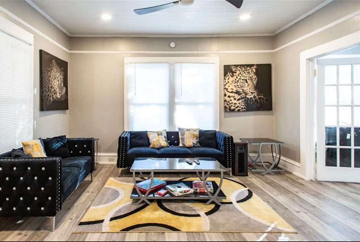 The living area features dark blue sofas with studded accents, complemented by a geometric area rug in shades of yellow and black. A coffee table holds magazines and decorative items, while large windows invite natural light, enhancing the spaciousness of the room.