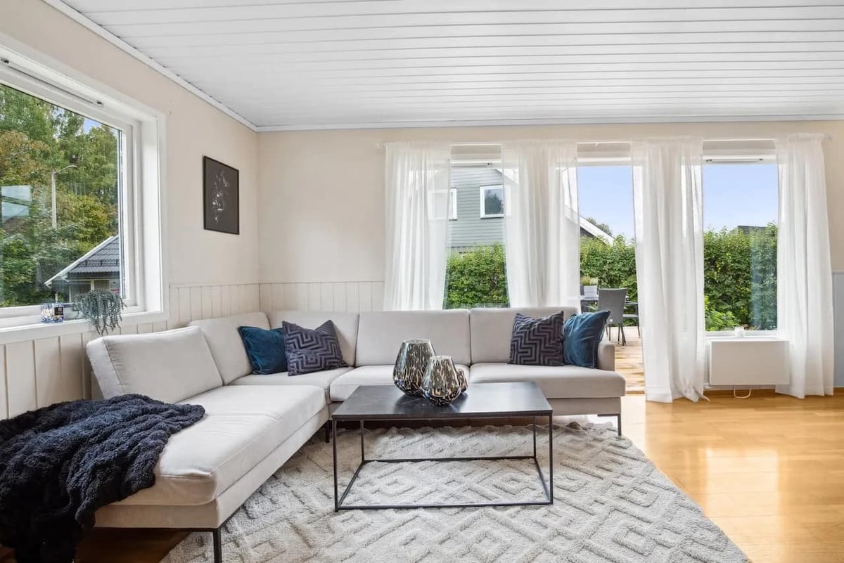 A bright living area features a spacious L-shaped sofa adorned with decorative cushions. A glass coffee table sits atop a textured area rug. Natural light streams through large windows, and sheer curtains gently frame the view of greenery outside.