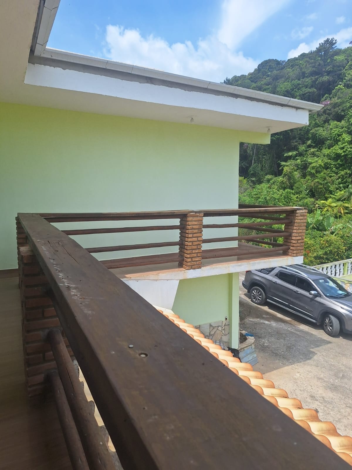 A wooden railing surrounds a balcony overlooking lush greenery. The light green exterior wall complements the natural surroundings, while a vehicle is parked on the driveway below. Bright blue skies and scattered clouds create a serene backdrop.