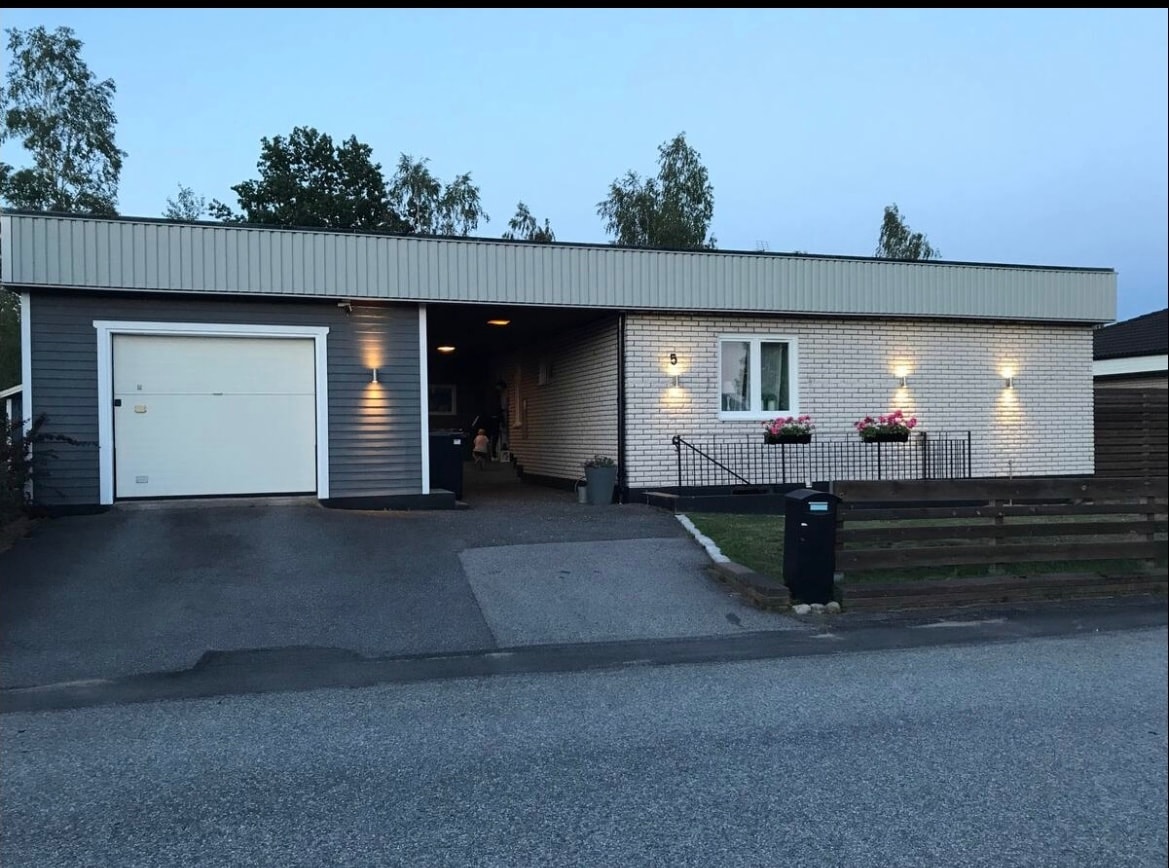 The exterior of a modern single-story house is presented, featuring a light brick façade and a dark gray accent wall. A driveway leads to a garage, and wall-mounted lights illuminate the entrance. Flower boxes adorn the front windows, enhancing the inviting appearance.