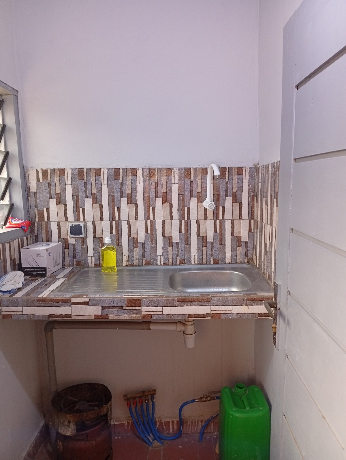 A compact kitchenette is featured, showcasing a stainless steel sink set against a tiled backsplash with a mix of neutral tones. A small window allows natural light to brighten the space. Cleaning supplies and plumbing fixtures are visibly organized along the wall.