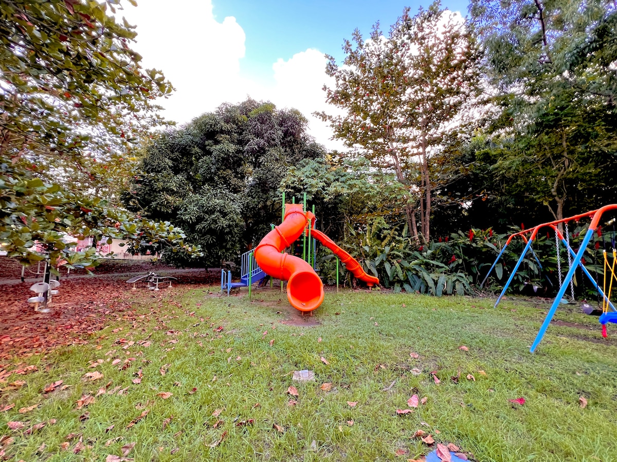 A vibrant children's playground is shown, featuring a large orange slide among lush greenery. Adjacent swings and climbing equipment are visible, surrounded by well-kept grass. The bright colors of the playground equipment complement the natural environment, creating an inviting space for outdoor play.