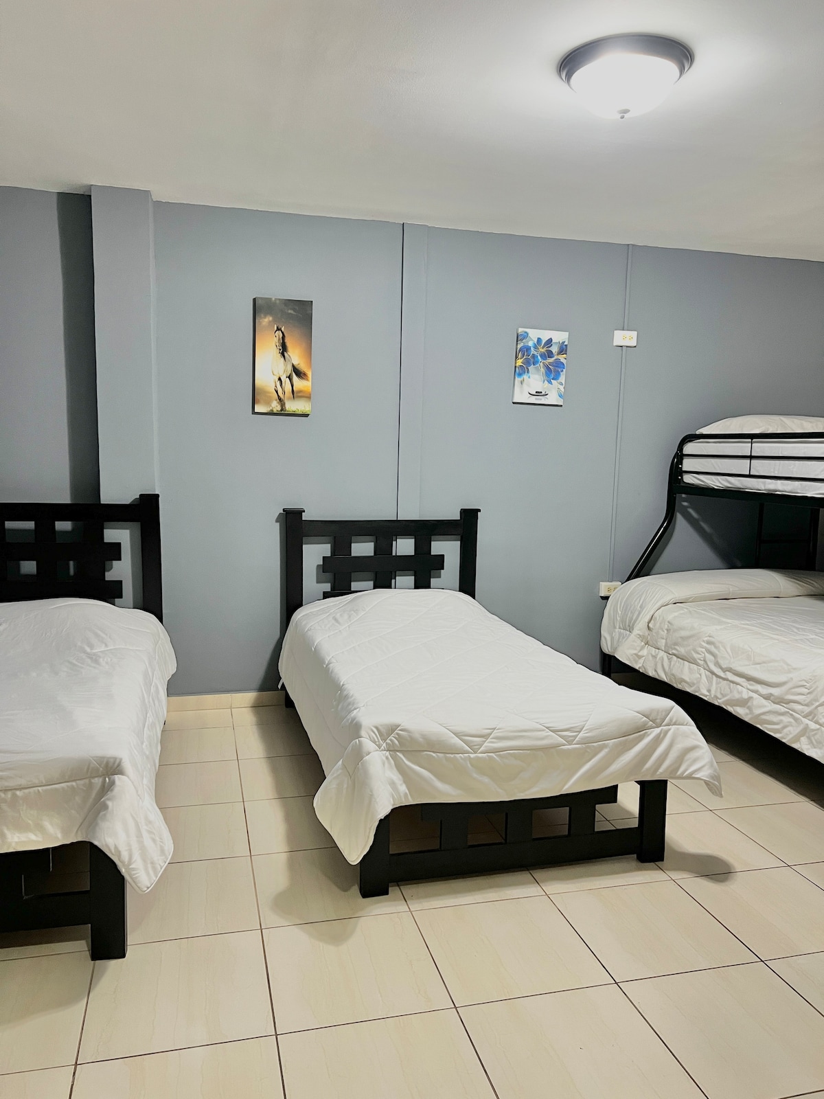 A simple sleeping area features three beds with clean, white bedding. The walls are painted in a calming light gray shade. Two pieces of artwork are hung on the wall above the beds, adding a touch of color to the space.
