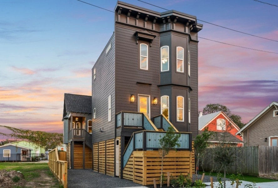 A contemporary two-story home with a striking dark exterior is presented. Large windows provide ample natural light, while the entryway is highlighted by elegant lighting fixtures. Surrounding greenery and a gravel path enhance the inviting exterior, creating a warm welcome for guests.