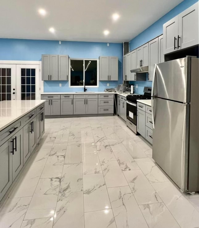 A modern kitchen is equipped with grey cabinets and stainless steel appliances, including a refrigerator and stove. A large island with white countertops provides ample workspace. Bright blue walls contrast with the light marble floor, enhancing the spacious feel of the area.