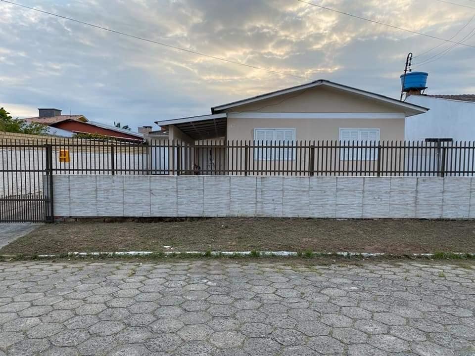 A single-story house is shown behind a low, textured wall. The front yard features a gravel surface, and a metal gate is visible. Large windows span the front of the home, allowing for ample natural light. The sky is overcast, casting a soft light over the scene.