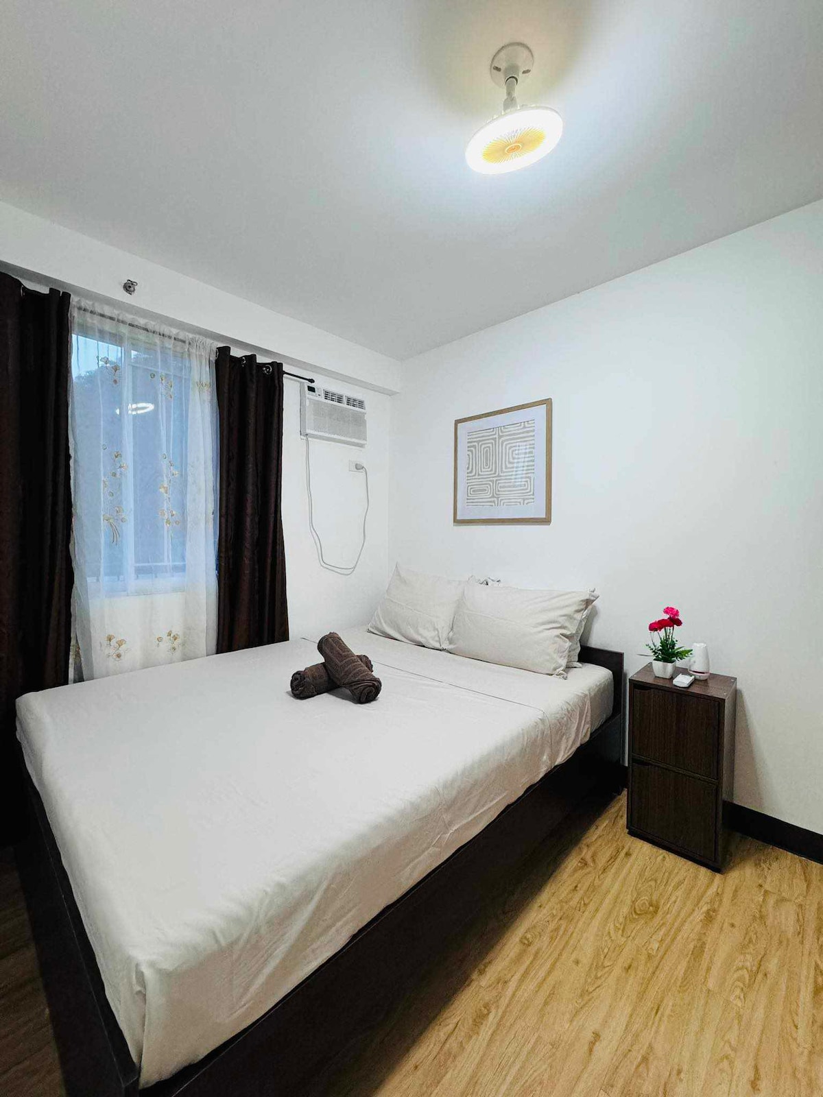 A cozy bedroom features a spacious bed with neutral bedding positioned against a white wall. Dark curtains frame a window, allowing natural light to enhance the space. A small bedside table holds a flower vase, adding a touch of color to the room.