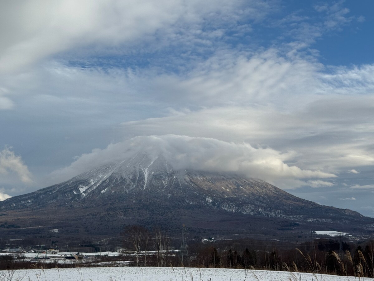 A snow-covered mountain is captured under a sky filled with soft clouds. The white landscape contrasts with the rugged mountain terrain, while the shadows hint at its majestic height. A serene atmosphere is conveyed through the gentle interplay of light and cloud formations.