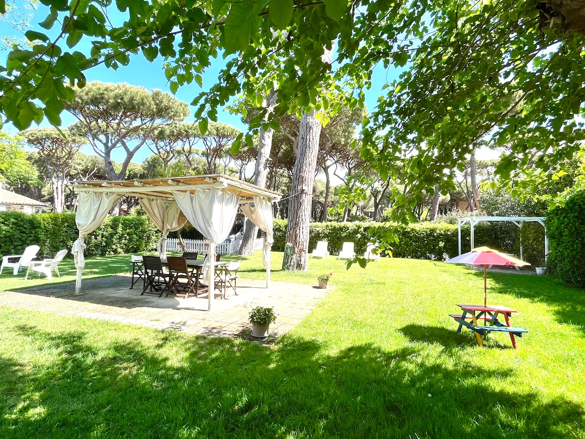 A well-maintained garden features a gazebo draped in light fabric, providing shade over a wooden dining table and chairs. Surrounding areas contain grassy spaces, a picnic table, and several lounge chairs, all under the canopy of tall pine trees.
