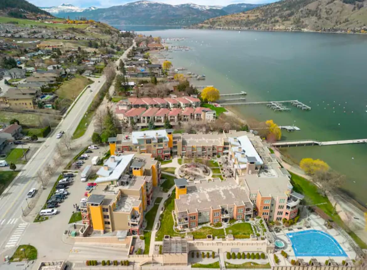 An aerial view displays the vibrant lakeside community, showcasing the arranged buildings around a central courtyard. The outdoor pool glistens under the sun, with lush greenery surrounding the property. Okanagan Lake and mountainous terrain provide a serene backdrop to the inviting lakeside amenities.
