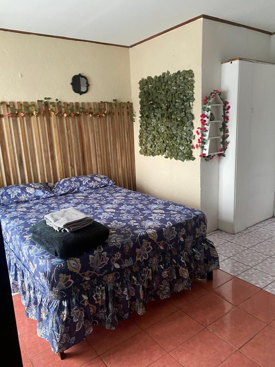 A sleeping area features a floral-patterned bedspread covering a spacious bed, complemented by neatly arranged towels. A wooden slatted headboard is adorned with greenery, while a white wardrobe stands nearby. Natural light enters through the window, enhancing the tranquil atmosphere.