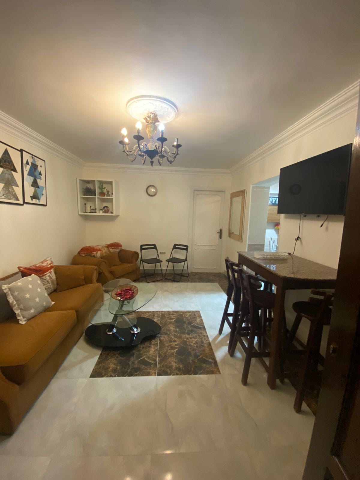 A living area is presented, featuring a comfortable brown sofa and a glass coffee table positioned on a marble floor. A small dining space with two chairs is visible, accompanied by a bar counter with stools. Decorative wall art and a chandelier add to the ambiance.