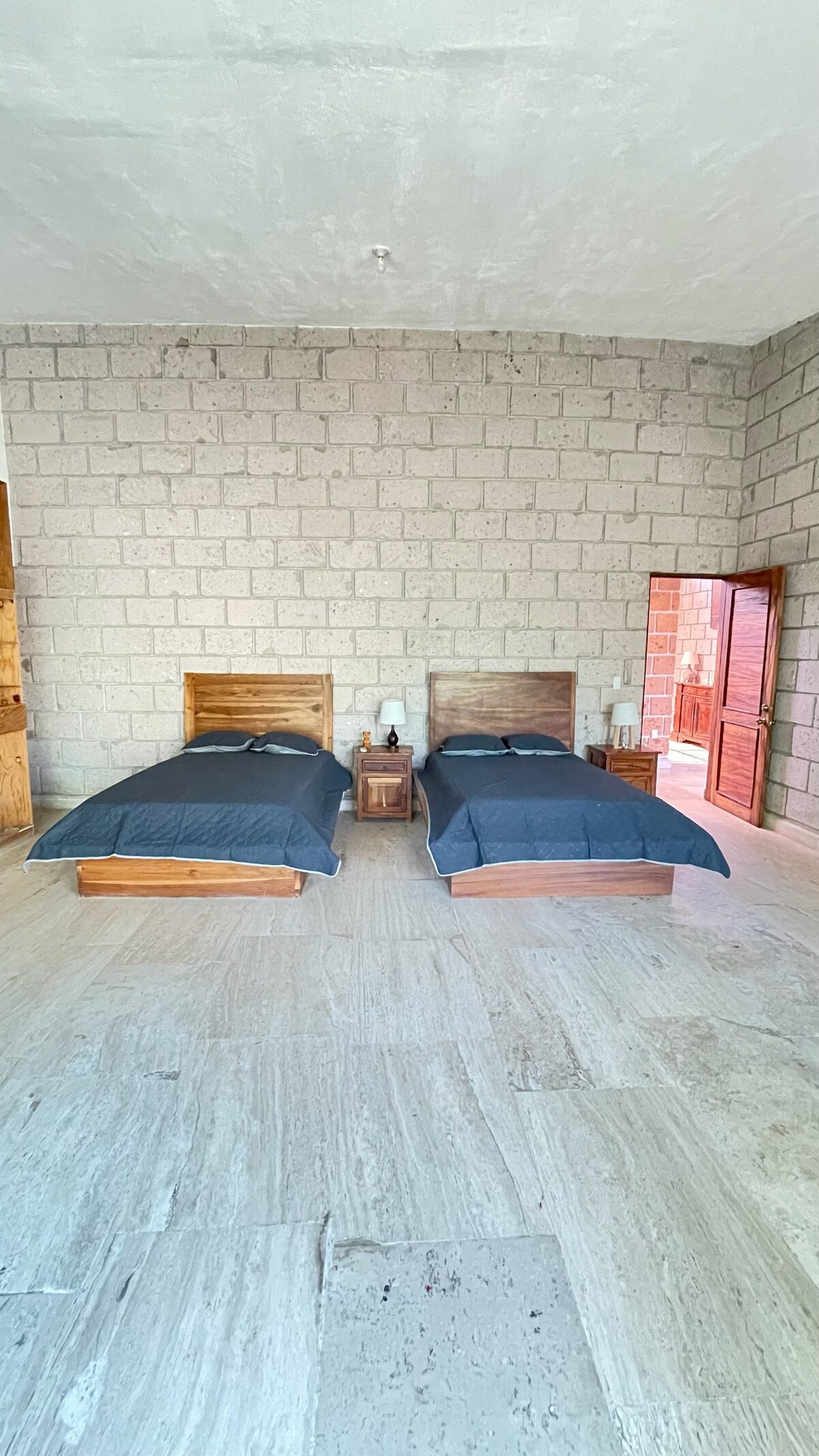 The room features two wooden beds with dark bedding set against a textured stone wall. Natural light enters through an open doorway, illuminating the light-colored flooring. Nightstands are positioned beside each bed, enhancing the space's functionality and comfort.