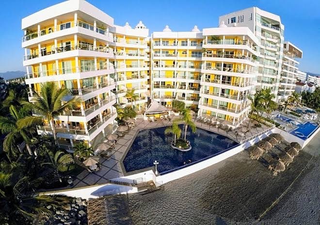 The building showcases a modern design featuring multiple stories with large balconies. A heated pool is surrounded by lounge chairs and tropical palm trees, with a glimpse of the sandy beach in the foreground. The clear blue sky enhances the resort-like ambiance.