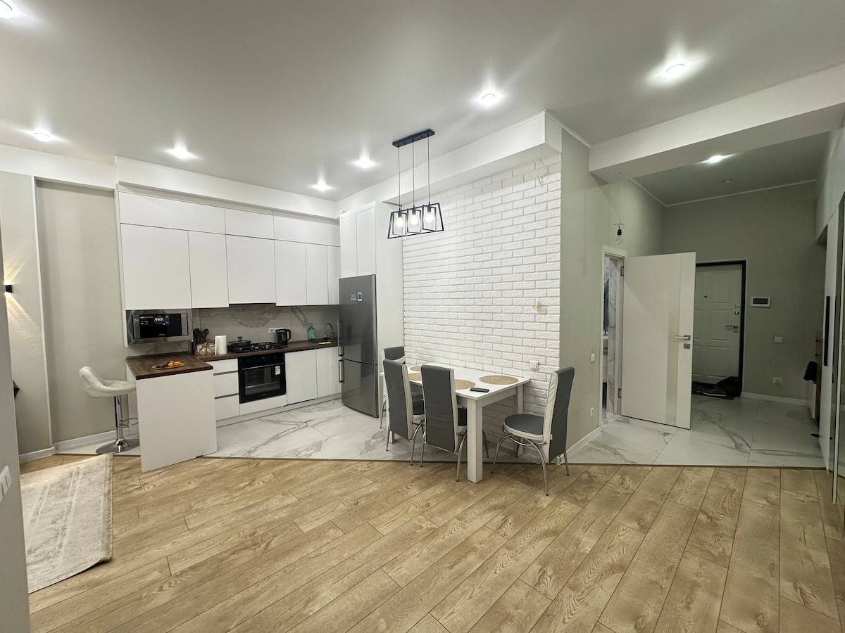 An open-plan living area is showcased, featuring a modern kitchen equipped with a refrigerator and cooking appliances. Dining space with a table and chairs is visible, and light-colored wood flooring complements the overall design. Soft lighting and neutral tones create a bright atmosphere.