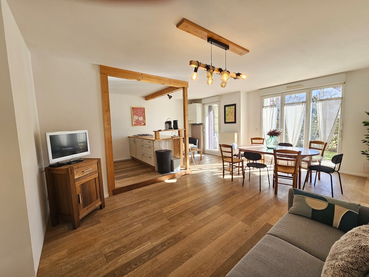 A spacious living area featuring wooden flooring and natural light. A dining table with several chairs is positioned adjacent to a fully equipped kitchen. A television stand is visible, and a comfortable sofa adds to the welcoming environment. Large windows provide a view of the outdoors.