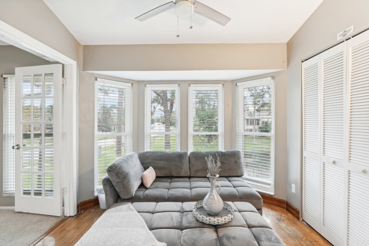 A bright and spacious living area features a large sectional sofa and a decorative coffee table. Multiple windows allow natural light to fill the room, while a ceiling fan adds comfort. A closet with sliding doors is visible, enhancing the overall functionality of the space.