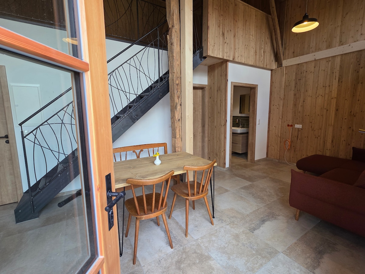 A dining area features a wooden table with four chairs, placed near a large window. A staircase with a decorative railing leads to an upper level. The walls are finished in natural wood, complemented by neutral tile flooring.