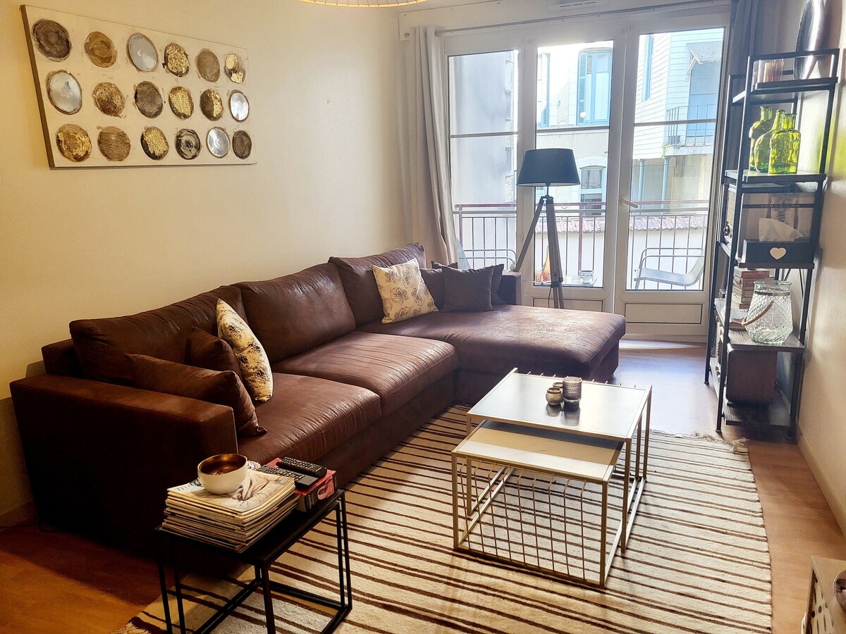 The spacious living area features a large brown sectional sofa, complemented by decorative pillows. A simple coffee table is centered on a patterned rug. Natural light floods the space through large glass doors, which lead to a balcony. Decorative items are placed on a shelving unit.