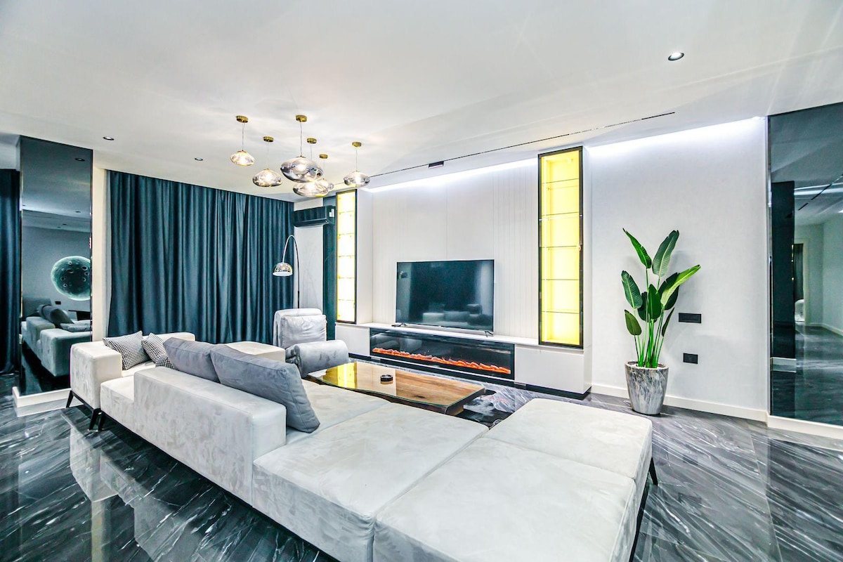 A modern living room features a large gray sectional sofa arranged around a glossy coffee table. A flat-screen television is mounted on the wall, with a contemporary electric fireplace below it. Tall plants and coordinated drapery add a touch of greenery and softness to the space.