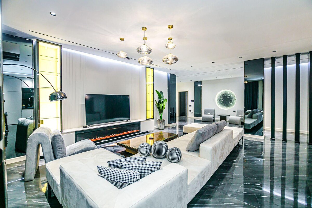 An elegant living area features a large sectional sofa adorned with decorative pillows and a sleek coffee table. A mounted television is positioned above a modern fireplace, while a decorative plant adds a touch of greenery. Bright lighting enhances the room's stylish design.