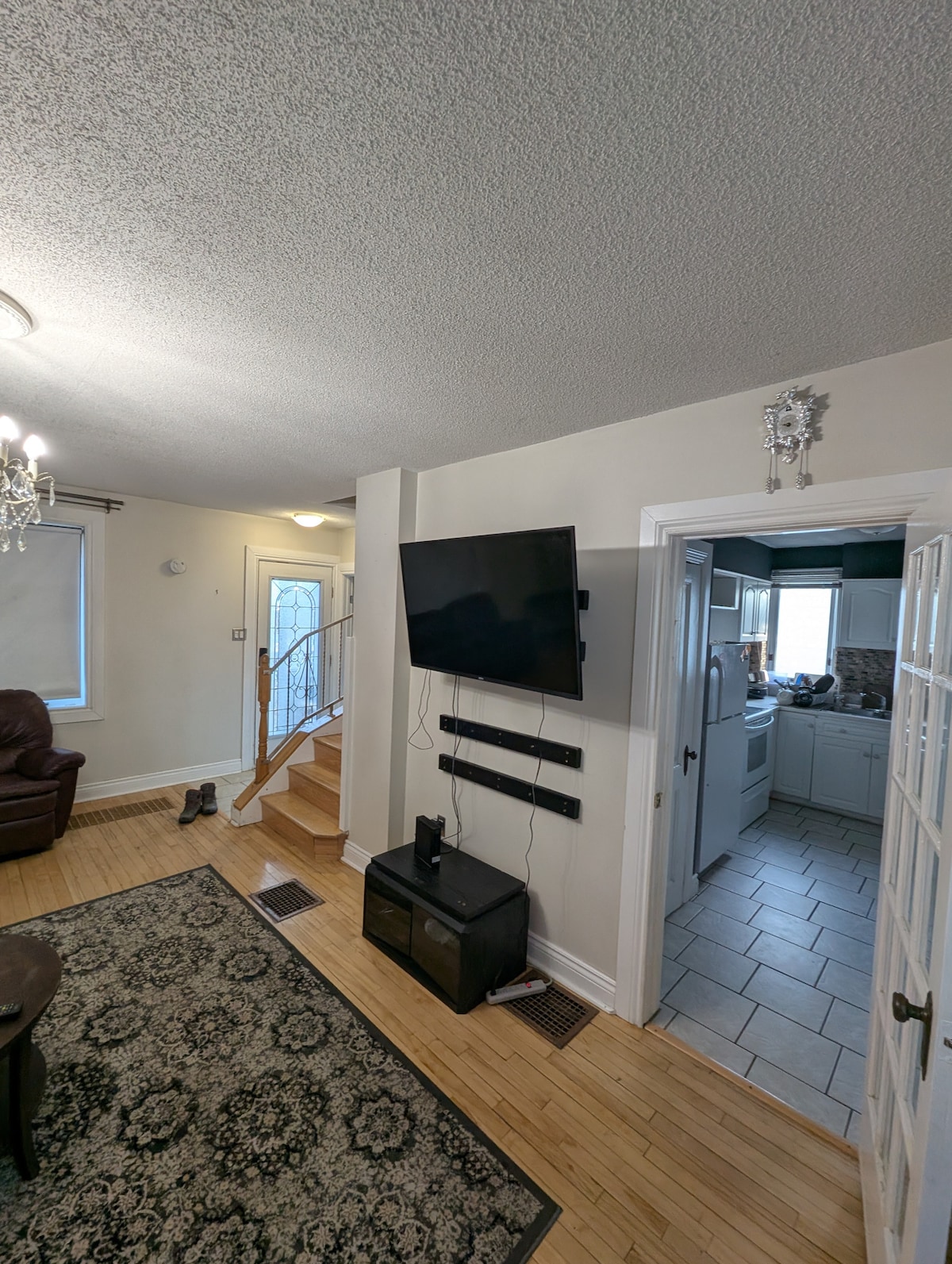 A living area is shown with hardwood flooring and a patterned area rug. A mounted television is positioned on the wall opposite a recliner chair. Doorways lead to a staircase on one side and a kitchen visible through an open door on the other.