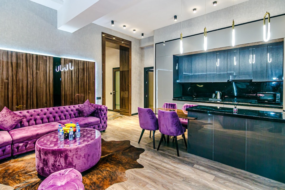 A stylish living area features a plush purple sofa and matching ottoman, complemented by sleek cabinetry and modern lighting fixtures. A dining set with purple chairs is positioned near the illuminated kitchen space, creating a modern and cohesive aesthetic throughout the room.